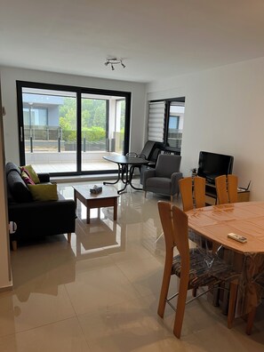 A modern living room with a sofa, a coffee table, a dining table with chairs, a TV, and a balcony with a view of greenery.