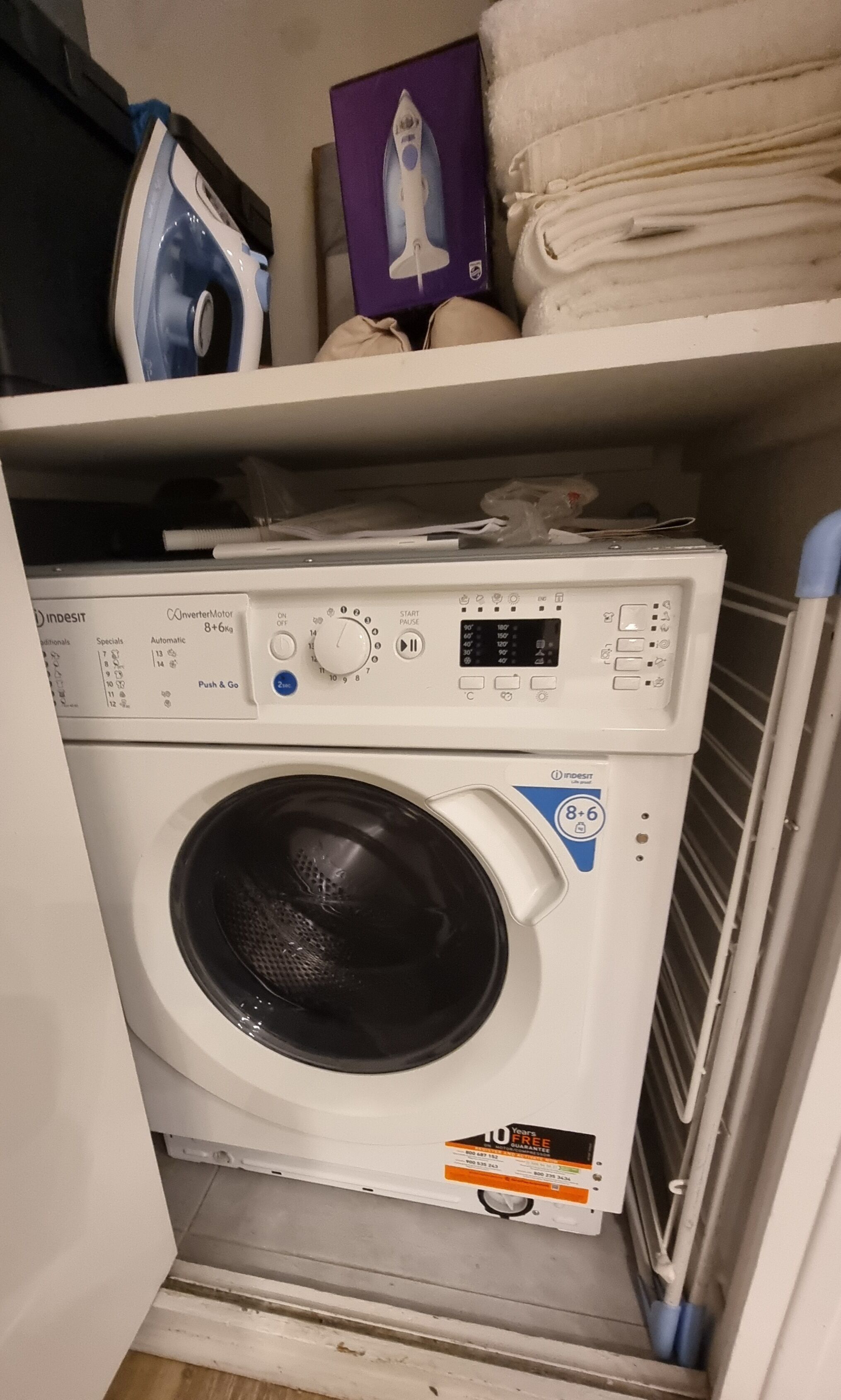 En kompakt tvättmaskin placerad i ett förråd, med en kontrollpanel, en etikett som anger en 10-årig garanti och en liten hylla ovanför där en bok och hopvikta handdukar förvaras.