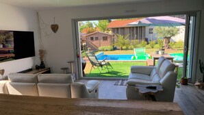 Ein modernes Wohnzimmer mit einem großen Schiebeglastür, die zu einem Garten mit Pool, Gartenmöbeln und Blick auf ein Haus führt.
