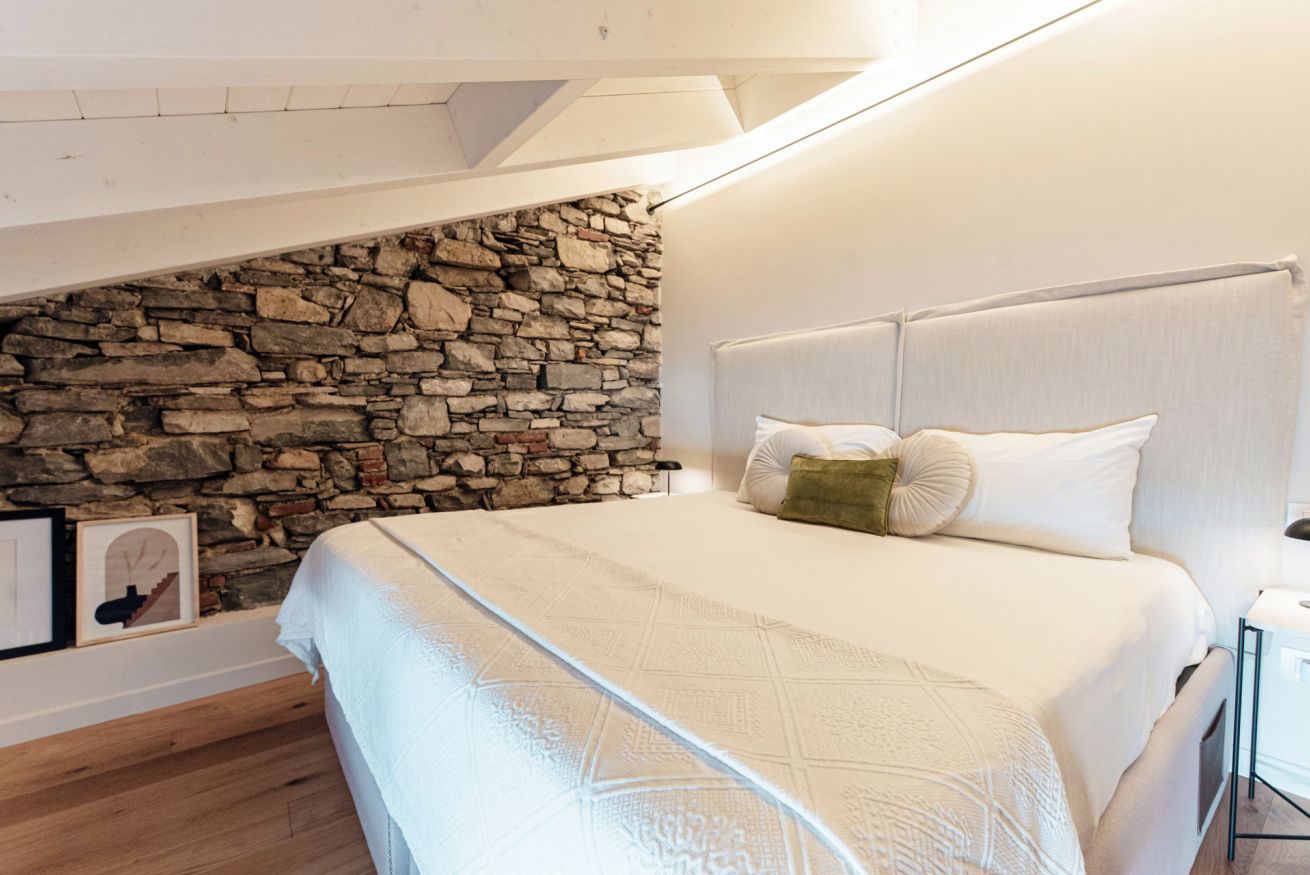 Quarto com parede de pedra, cama com roupa de cama branca e chão de madeira.