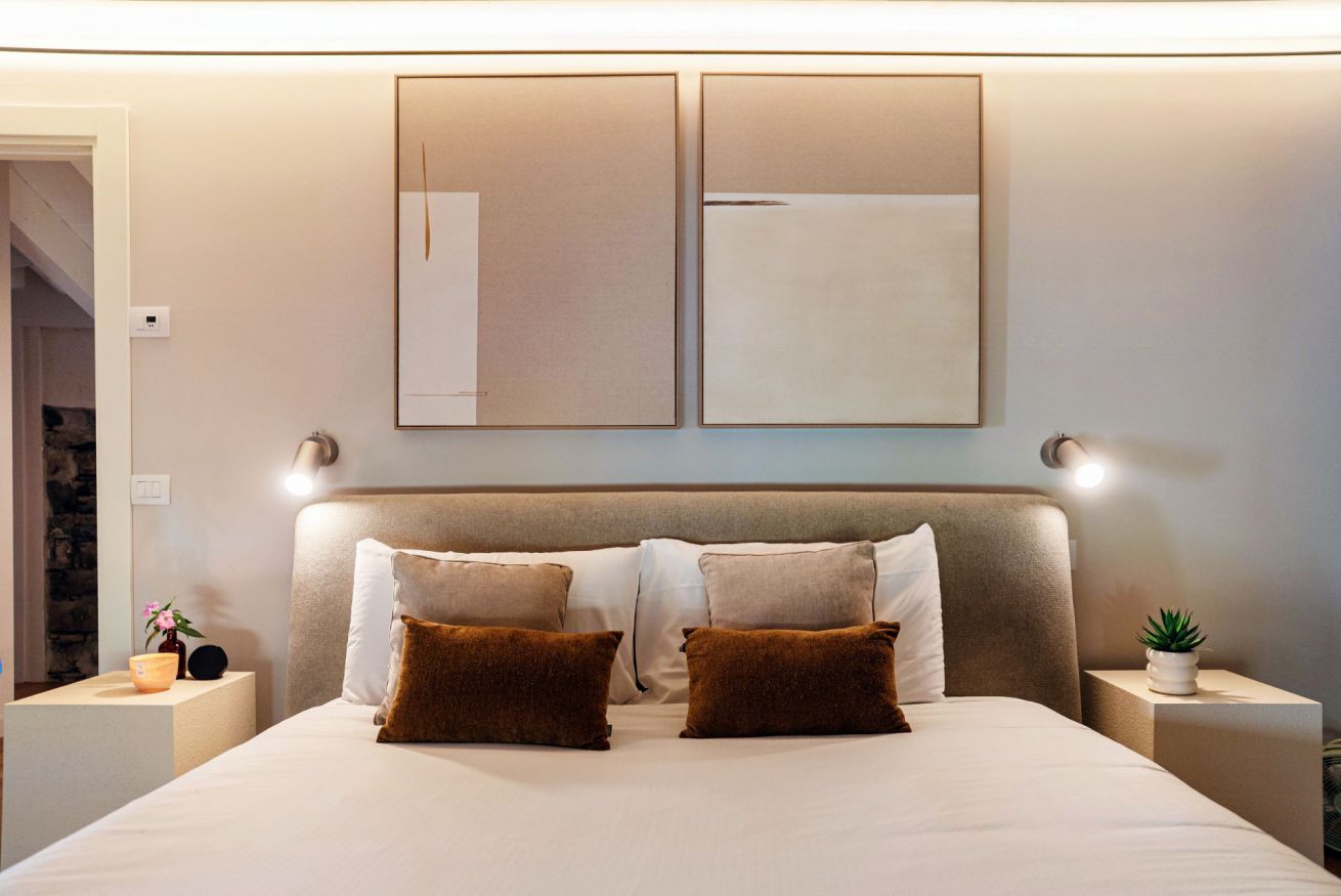 Cama bem arrumada com duas almofadas castanhas, cabeceira e duas luminárias fixadas na parede.