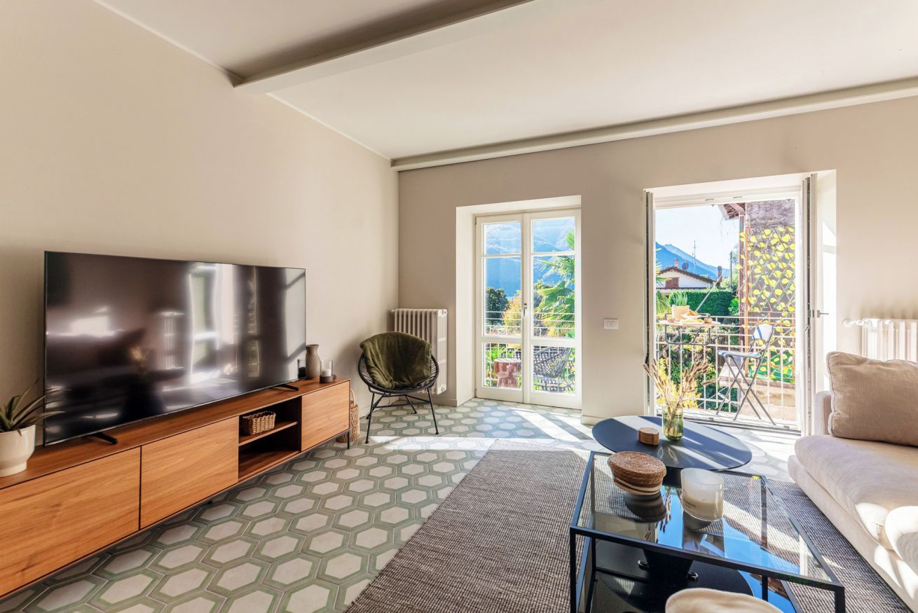 A living room with a large flat-screen TV, a sofa, a coffee table, and a view of a balcony with outdoor furniture.