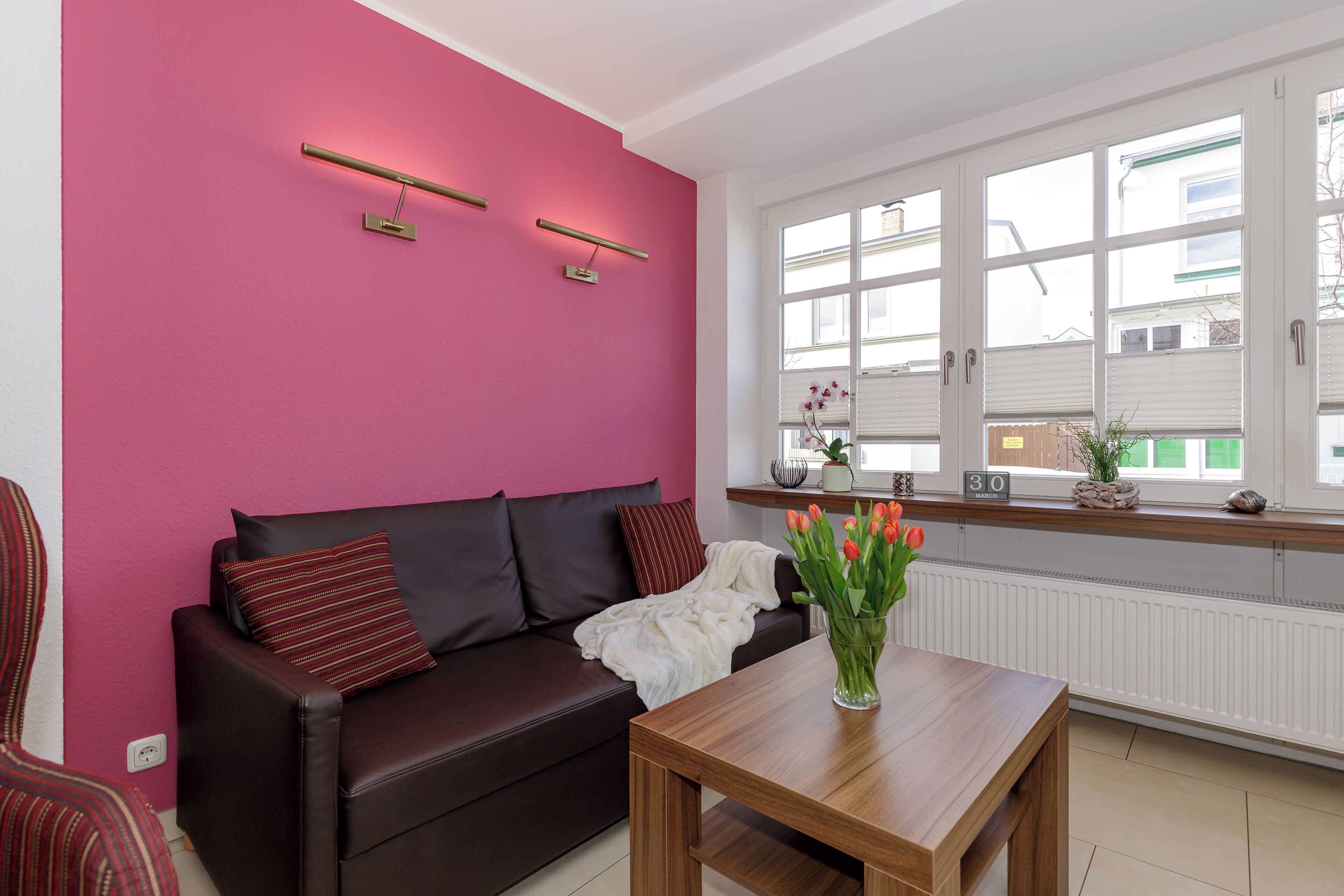 Een woonkamer met een bruine bank, een houten salontafel, een roze wand en een raam met zonwering.