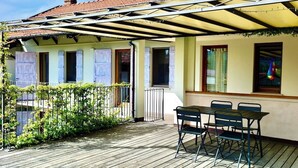 A covered outdoor patio with a table and chairs, a wooden deck, and a building with shutters. - Green retreat with sauna and terrace: authentic mini apartment in Magnano (Piemonte)