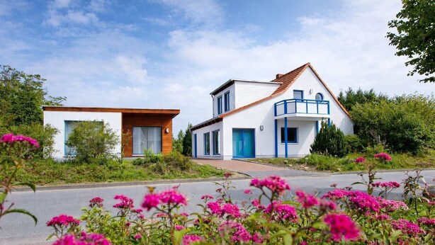 A modern two-story house with a white exterior and blue accents, surrounded by lush greenery and vibrant pink flowers.
