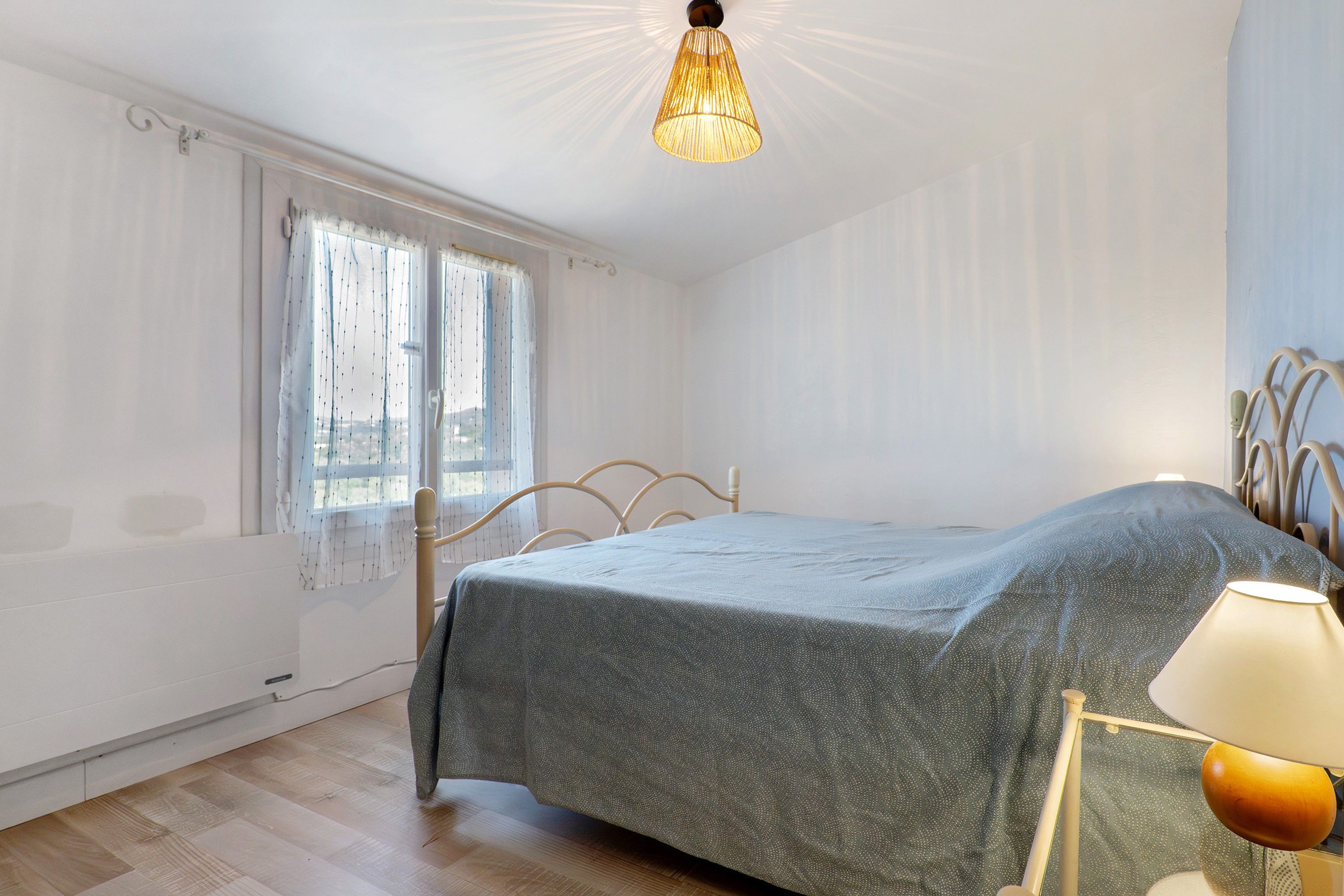 A bedroom with a bed, a window with curtains, a hanging light fixture, and a radiator.