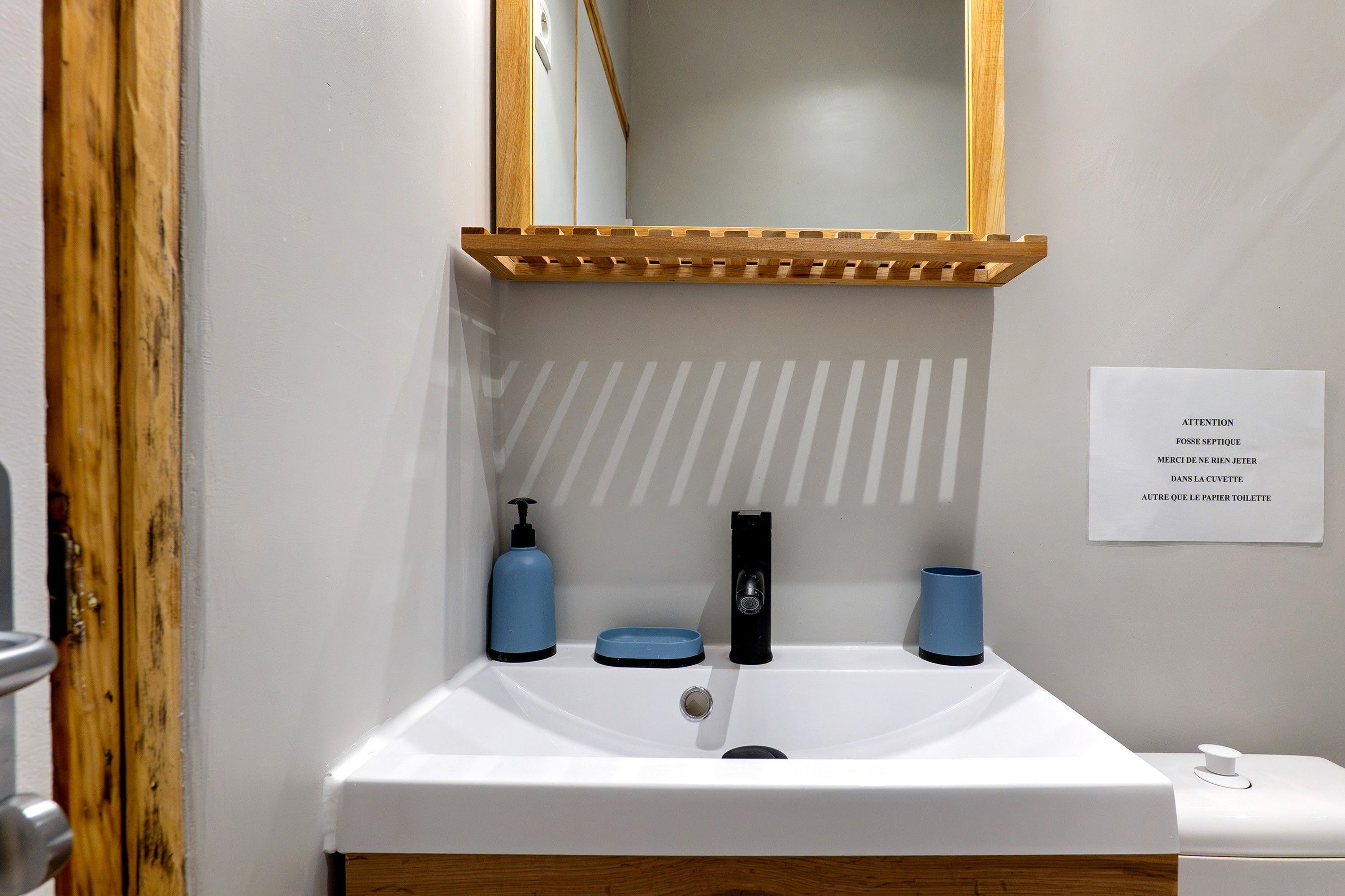 Un bagno con un lavandino bianco, uno specchio e una mensola in legno sopra. Si notano due erogatori azzurri e un rubinetto nero. Un cartello con del testo è visibile sulla parete.