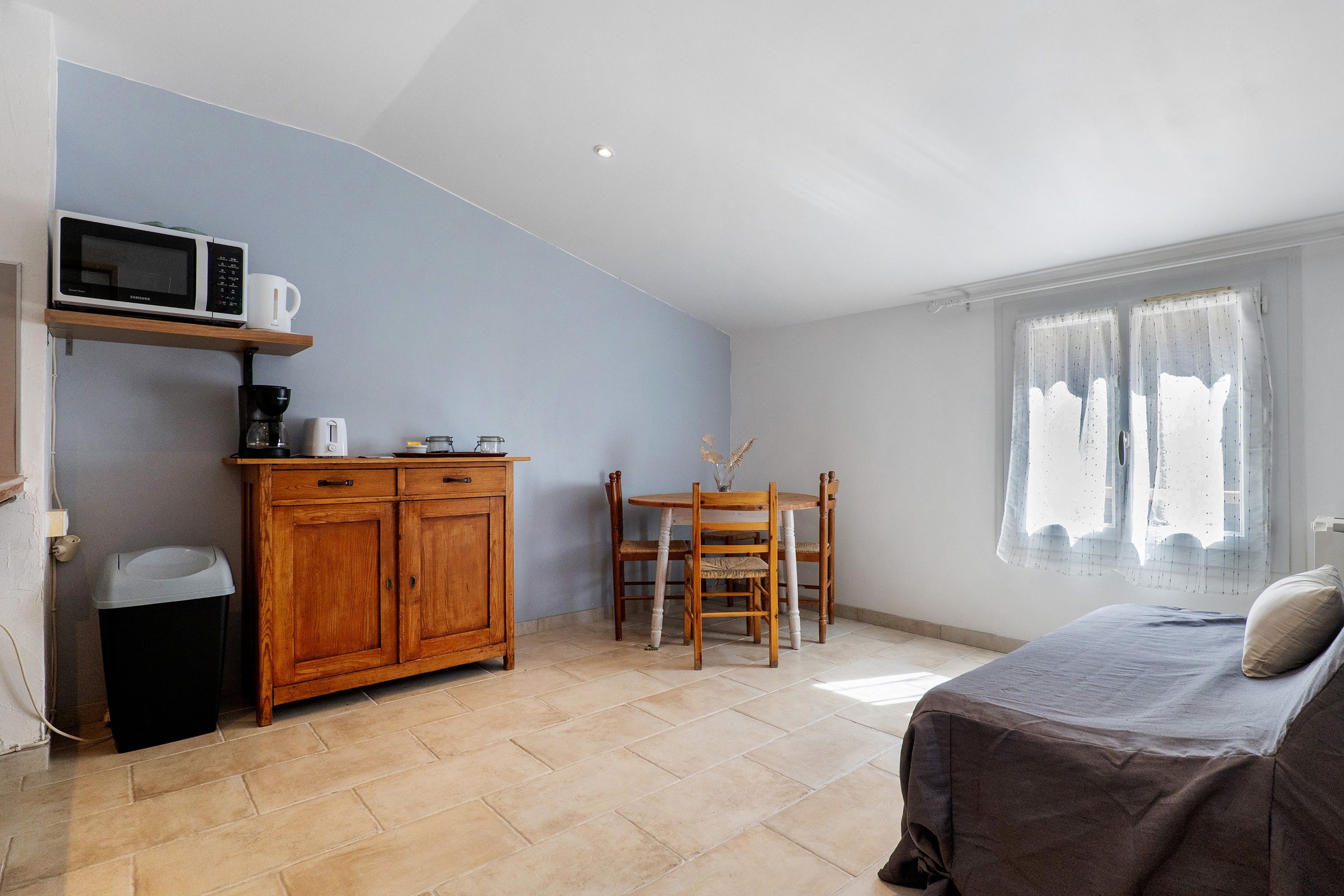 Quarto compacto com cama, um armário de madeira, uma pequena mesa com cadeiras e uma janela com cortinas.
