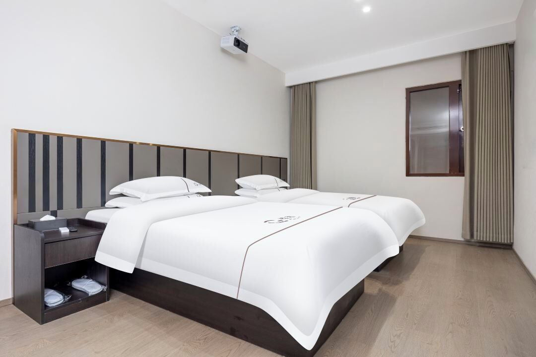 A modern hotel room with a large bed, a bedside table, a projector, and a window with curtains.