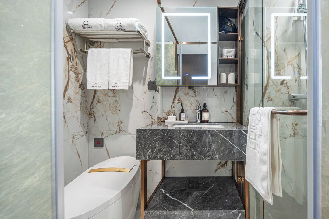 A modern bathroom with a marble wall, a glass shower enclosure, a white toilet, a vanity with a black marble countertop, and a mirror with integrated lighting.