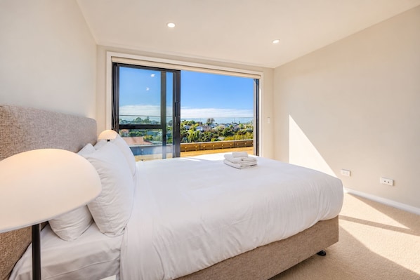 A bedroom with a large bed, a bedside table, and a view of a balcony with a cityscape. - Modern 4BR Coastal Stay /Sea Views & Walk to Beach (Torbay)