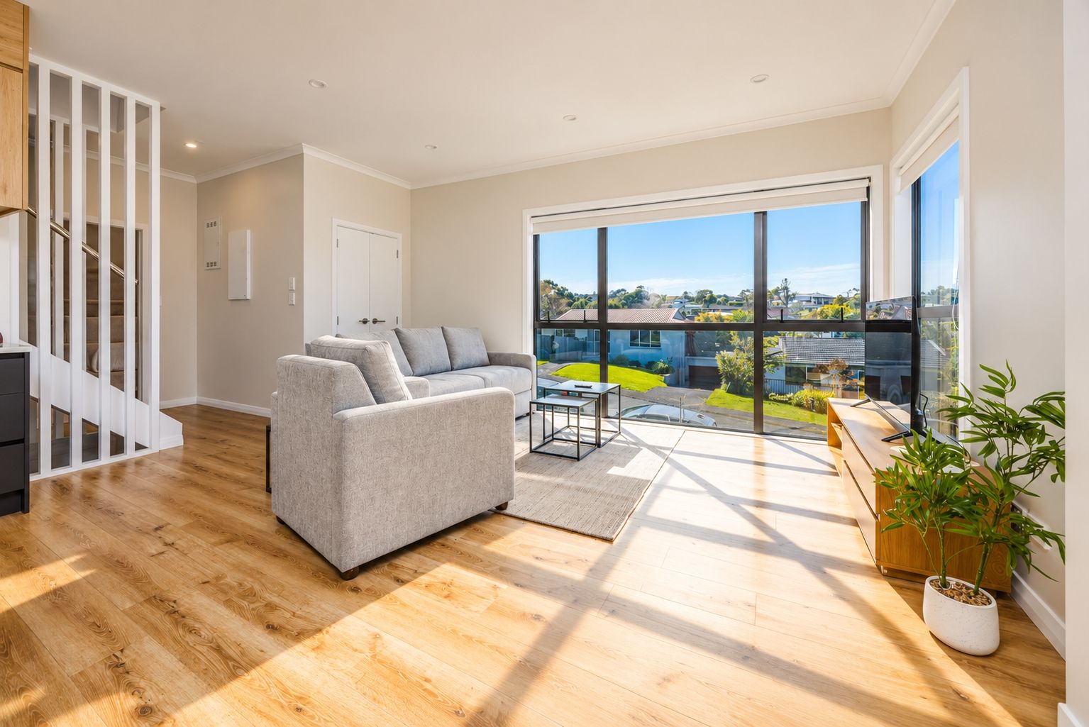 Ein modernes Wohnzimmer mit grauem Sofa, Holzboden, einem Esstisch und einer großen Glasschiebetür, die auf einen Balkon mit Blick auf die Nachbarschaft führt.