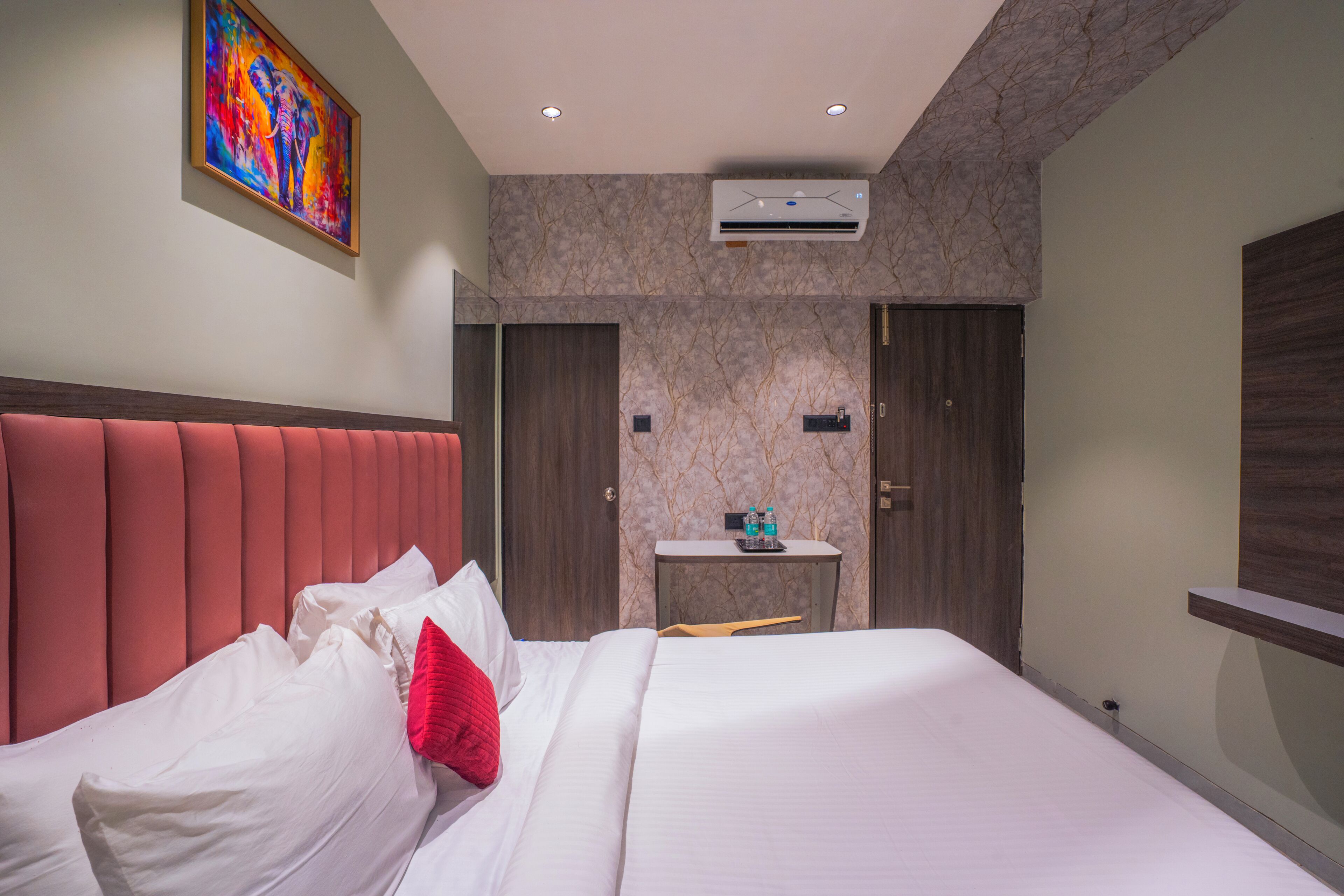 A hotel room with a bed, a headboard, a painting, an air conditioning unit, a small table with a mirror, and a door.