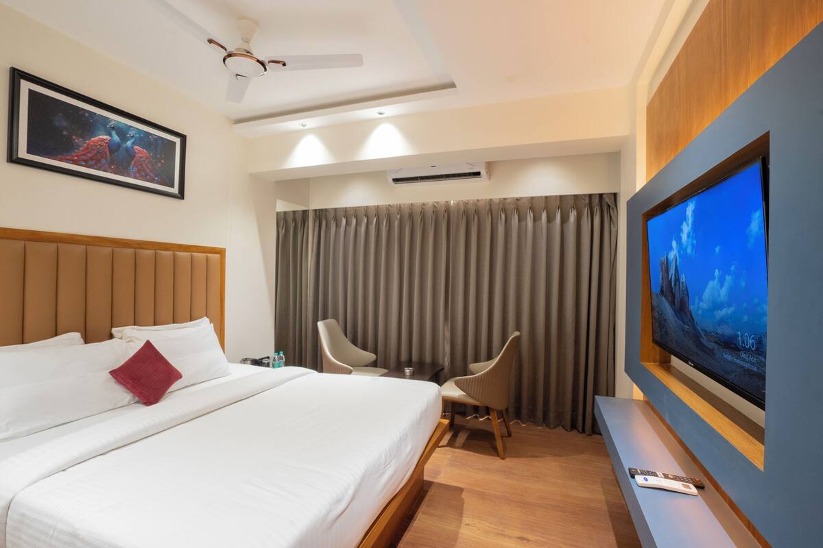 A hotel room with a bed, two chairs, a TV, and an air conditioning unit.