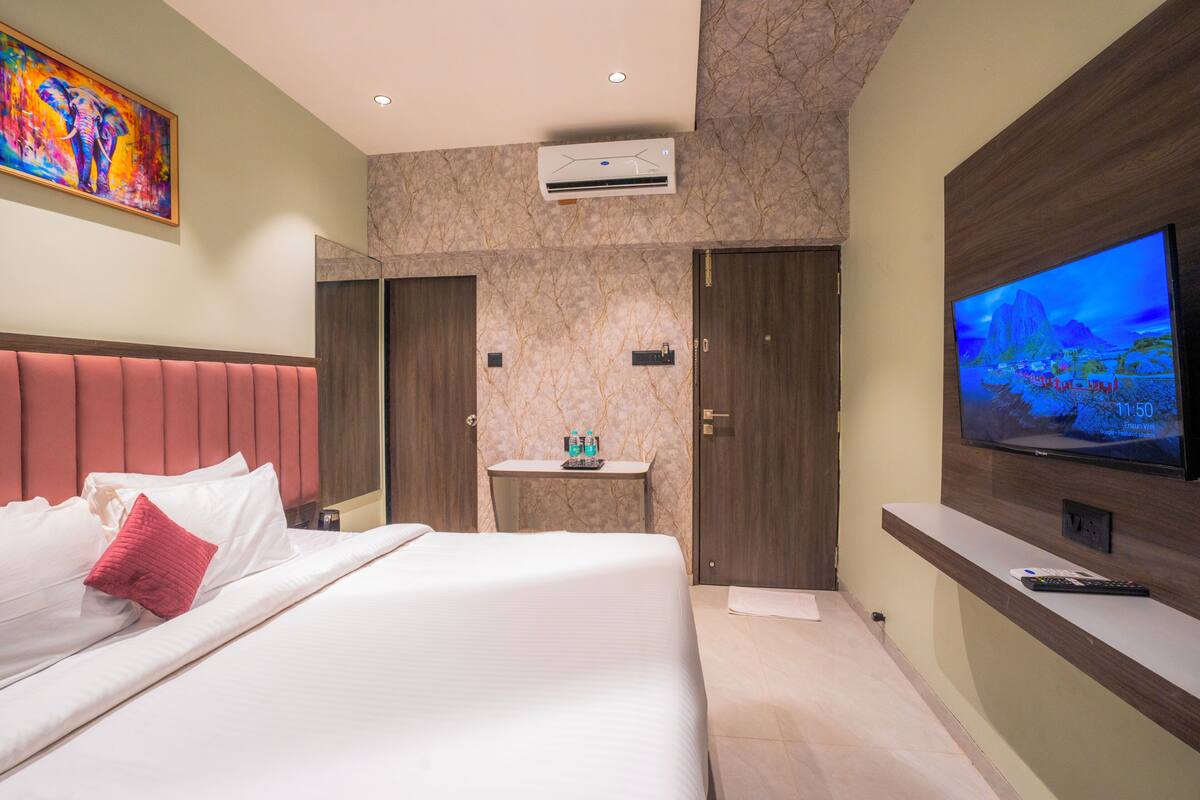 A hotel room with a bed, a TV, a small table with bottles, and a wall-mounted air conditioner.