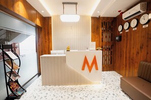 A reception desk with a large 'M' logo, a wall-mounted clock, and a bulletin board with magazines. - Mosaic Go Star Grand (Navi Mumbai)
