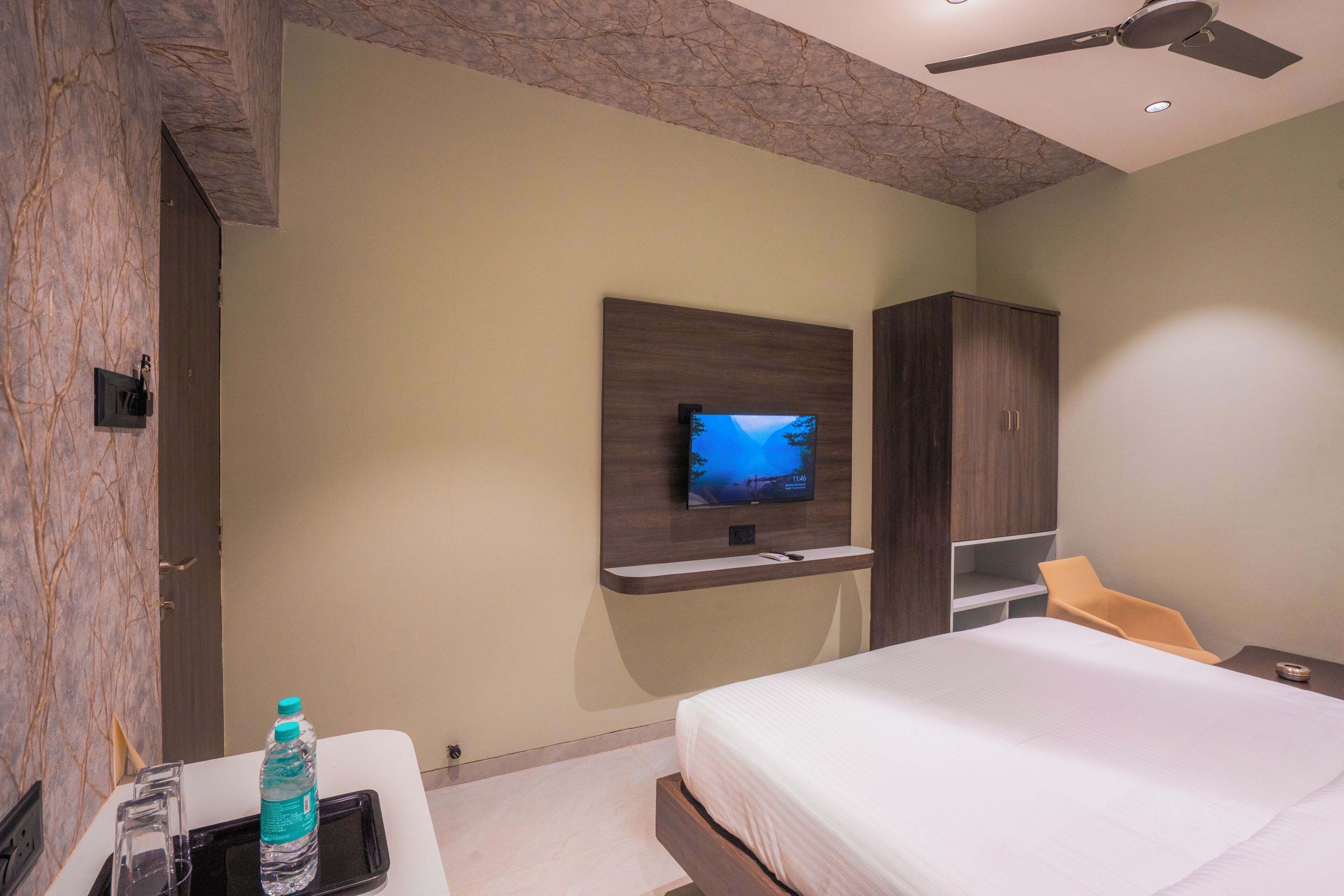 A modern hotel room with a bed, TV, wardrobe, and a small table with a water bottle.