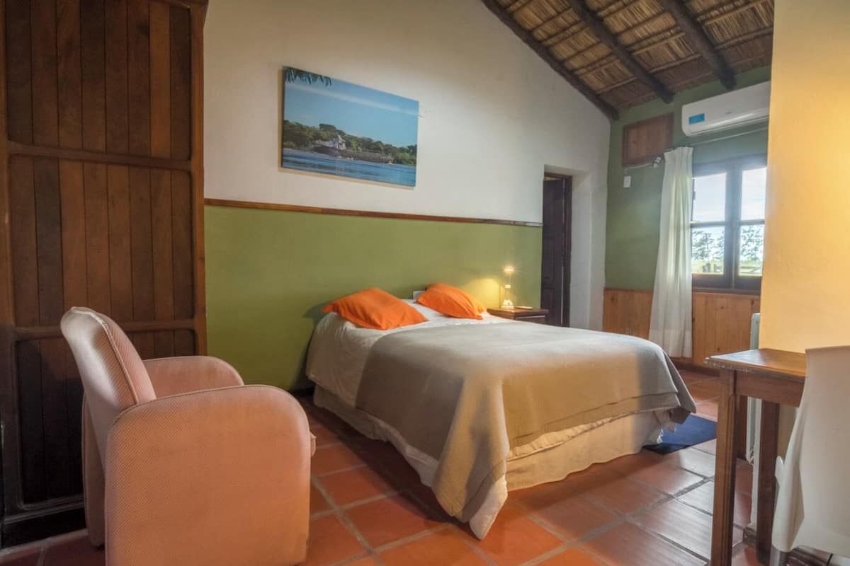 A bedroom with a bed, a chair, a nightstand, a window with curtains, and a painting on the wall.