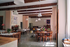 A spacious dining area with wooden tables and chairs, pendant lighting, and a view into another room with a fireplace. - La Magnanarié, a large family home in Provence (Villedieu)