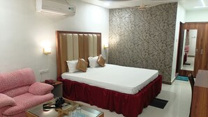 A hotel room with a bed, a red bed skirt, a pink sofa, a glass coffee table with a camera, and an air conditioning unit. - HOTEL VAIDYA REGENCY (Balaghat)