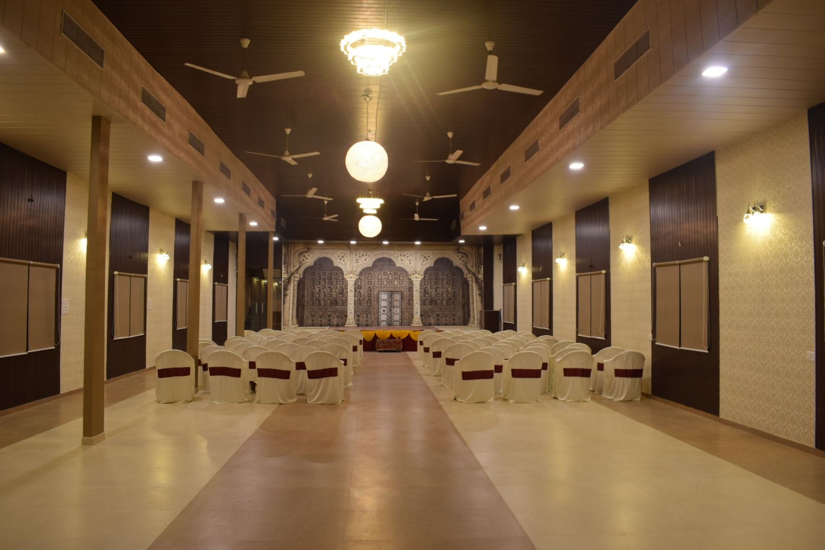 Un salón de conferencias con filas de sillas, un escenario con un telón de fondo, ventiladores de techo e iluminación empotrada.