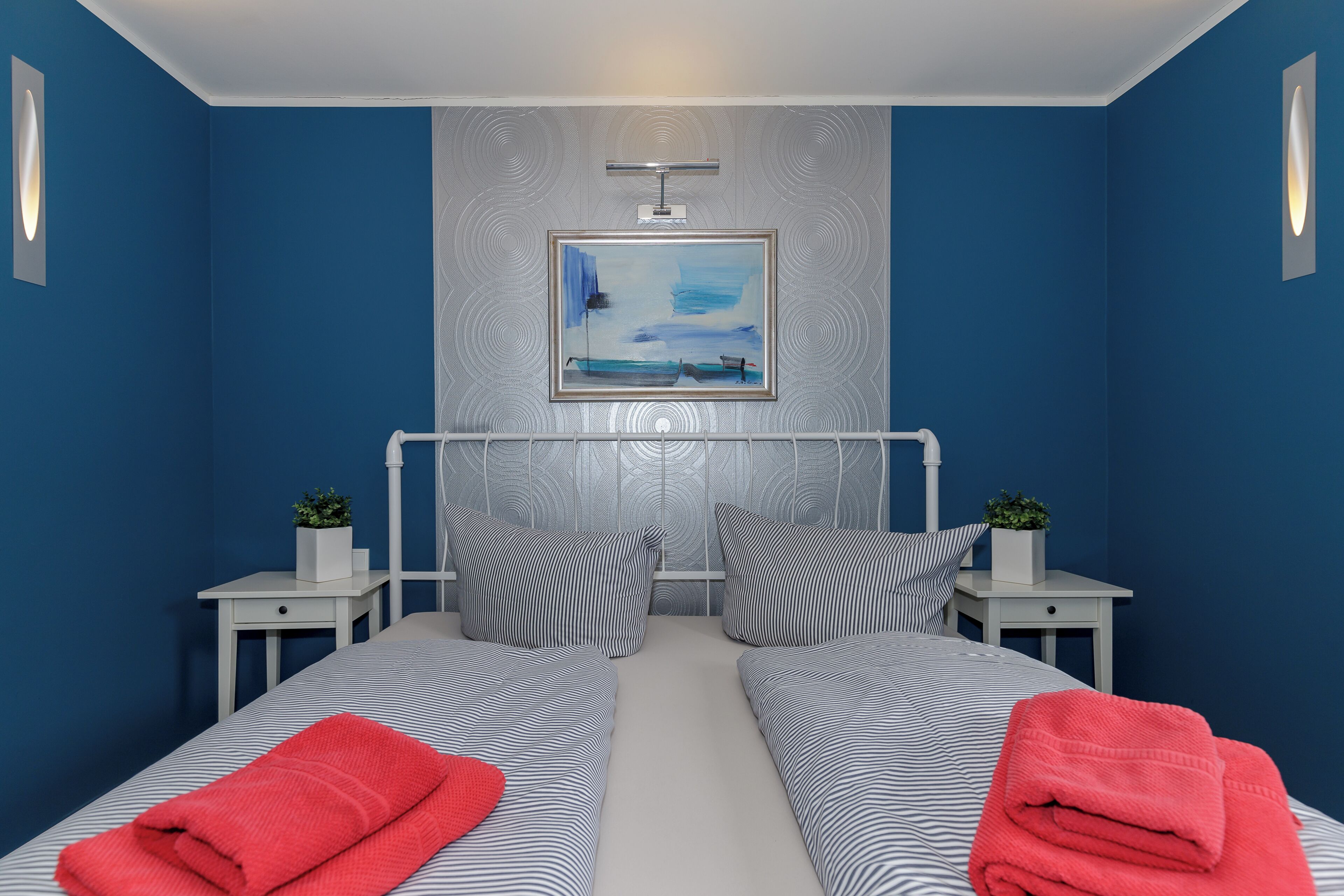 Une chambre avec deux lits, un tableau, un luminaire au plafond et deux tables de chevet ornées de plantes.