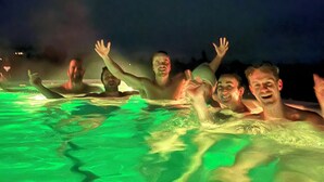Um grupo de pessoas em uma piscina, com os braços levantados, possivelmente em um resort ou hotel.