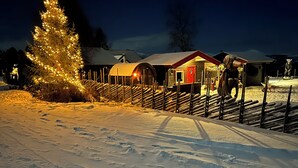 Paisagem coberta de neve com um pinheiro decorado para o Natal e um edifício rústico com uma porta vermelha.