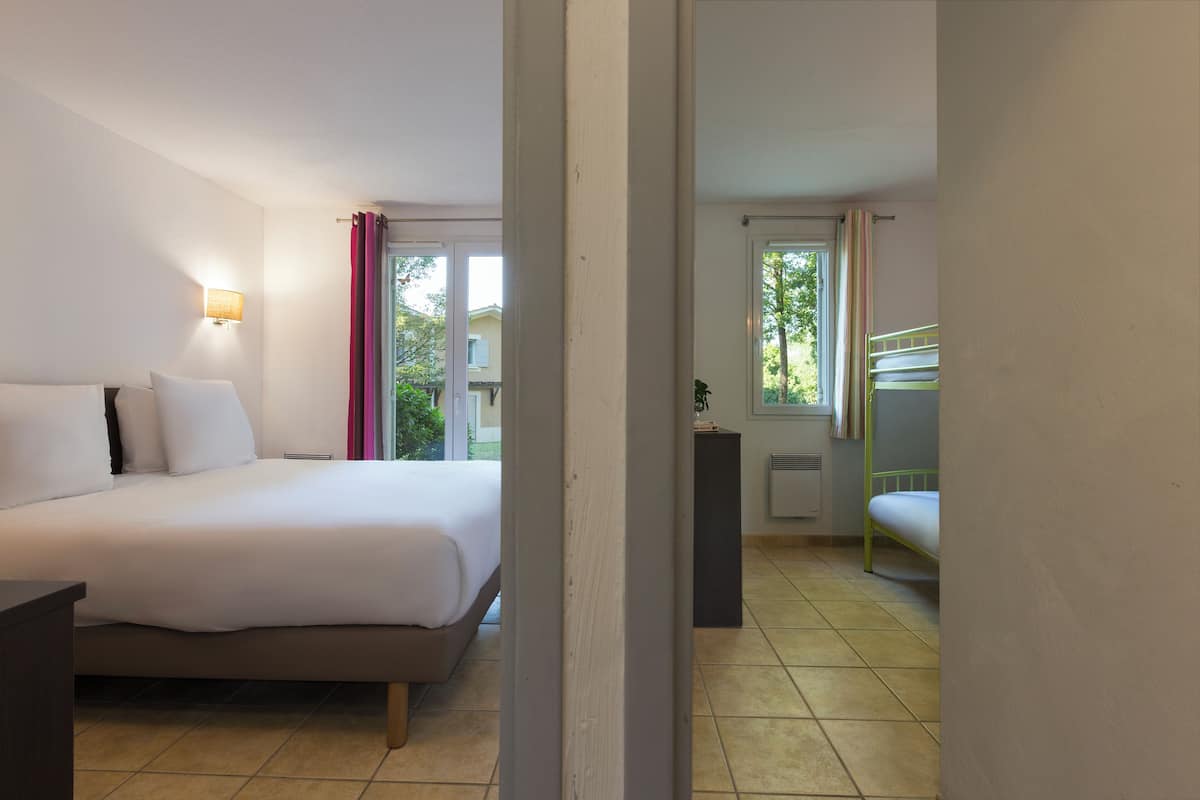 A hotel room with a bed, a bedside table, a lamp, a window with curtains, and a view of greenery outside.