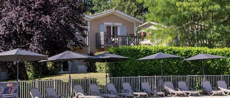 A swimming pool with lounge chairs and umbrellas, surrounded by greenery and a building in the background.