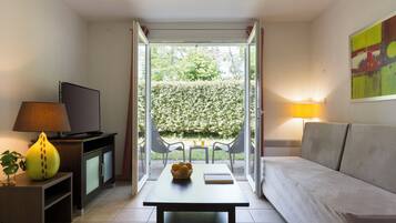 Un salon comprenant un canapé, un téléviseur, une table avec un panier de fruits, et donnant sur un jardin grâce à une porte vitrée.