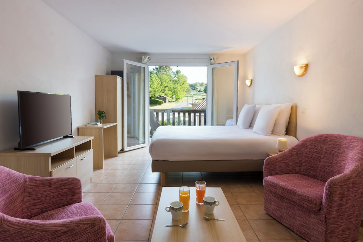 A hotel room with a bed, two pink armchairs, a TV, a coffee table with two cups and a glass, and a balcony with a view.