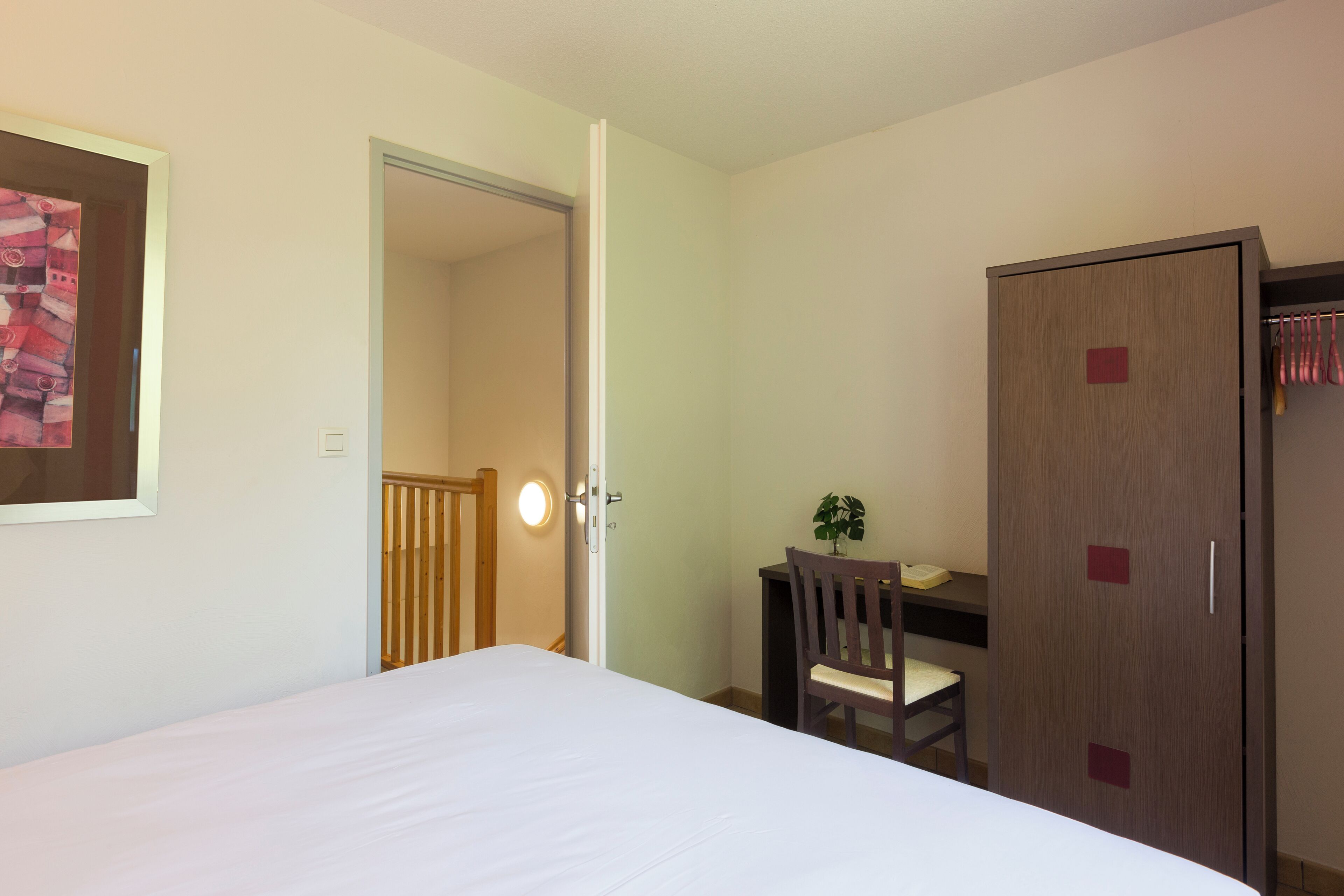 A hotel room with a bed, a desk with a chair, a wardrobe, and a door leading to another room.