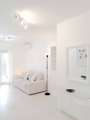 Ein weißes Zimmer mit einer Couch, einer Stehlampe, einem Glastisch und einem Fenster mit einer Pflanze.