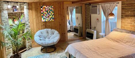 A cozy room with a bed, a chair, a potted plant, a colorful wall art, and a mirror.