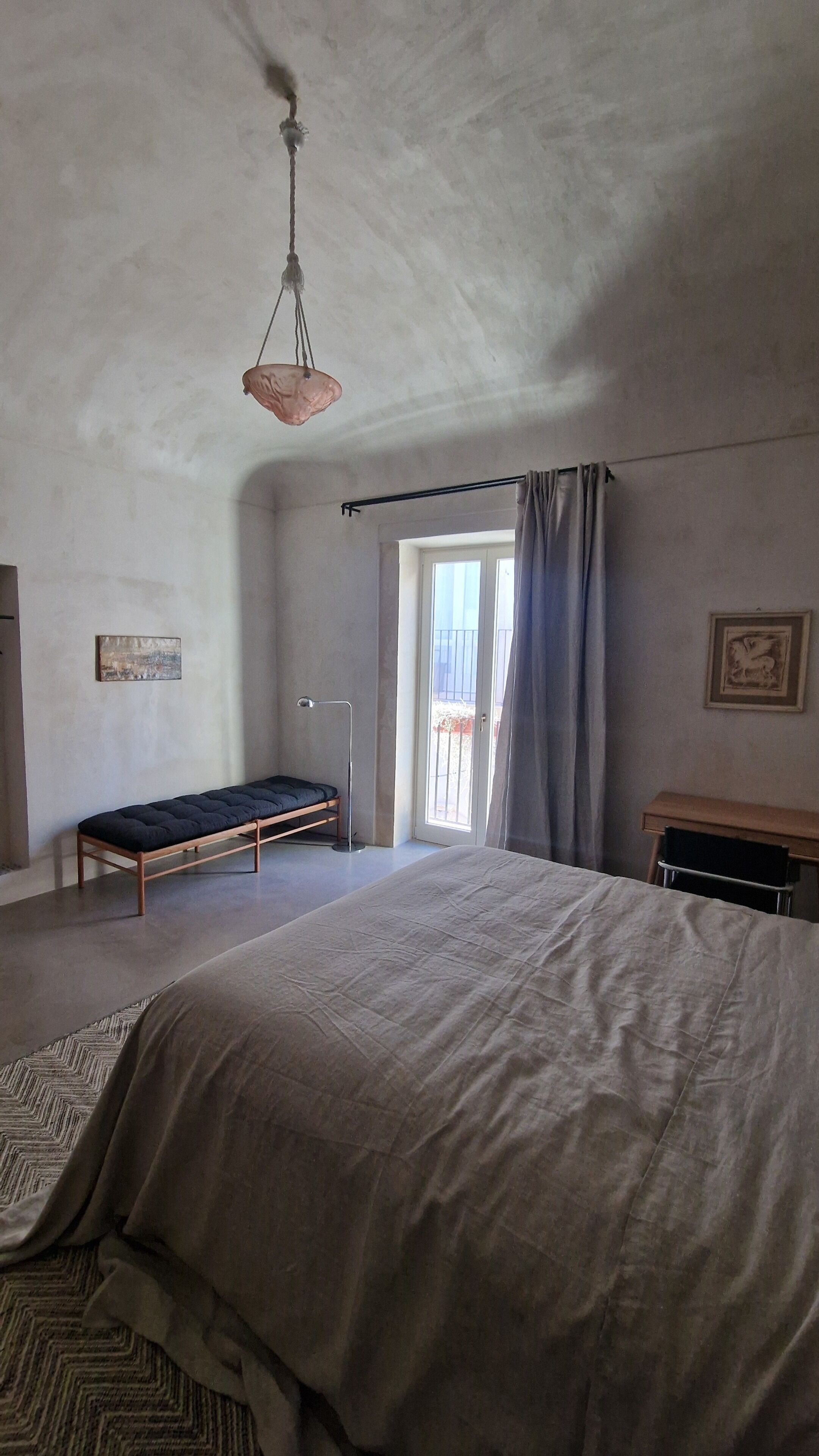 Un dormitorio con cama, una banca, un escritorio, una lámpara colgante y una puerta que da a un balcón.