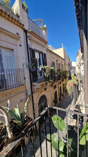 A narrow street with buildings on either side, featuring balconies with plants, a wrought iron gate, and a cobblestone path. - Casa Lureca (Syracuse)