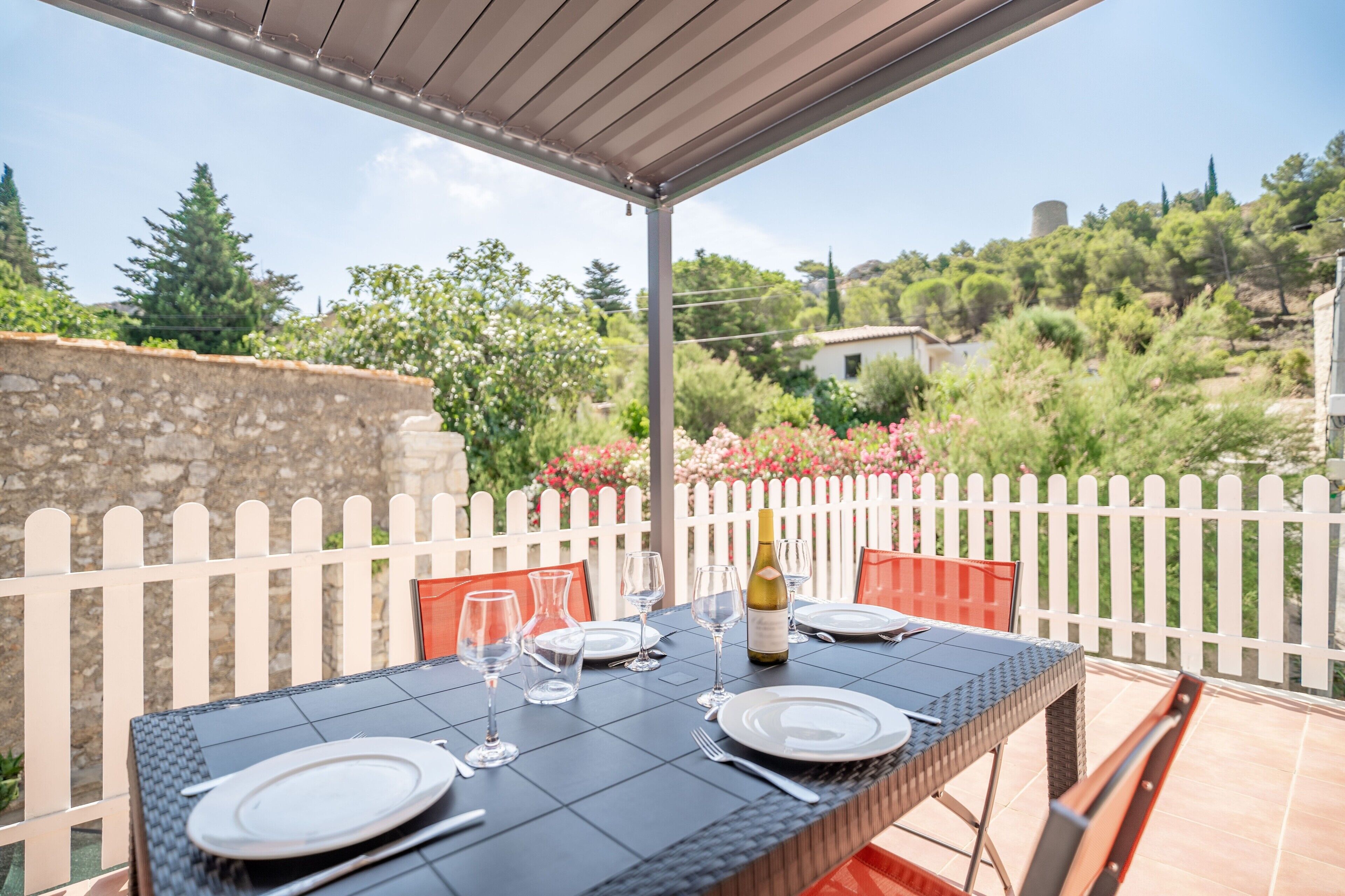 Un espacio de comedor al aire libre con una mesa dispuesta para dos personas, una botella de vino y una cerca blanca de piquetes al fondo.