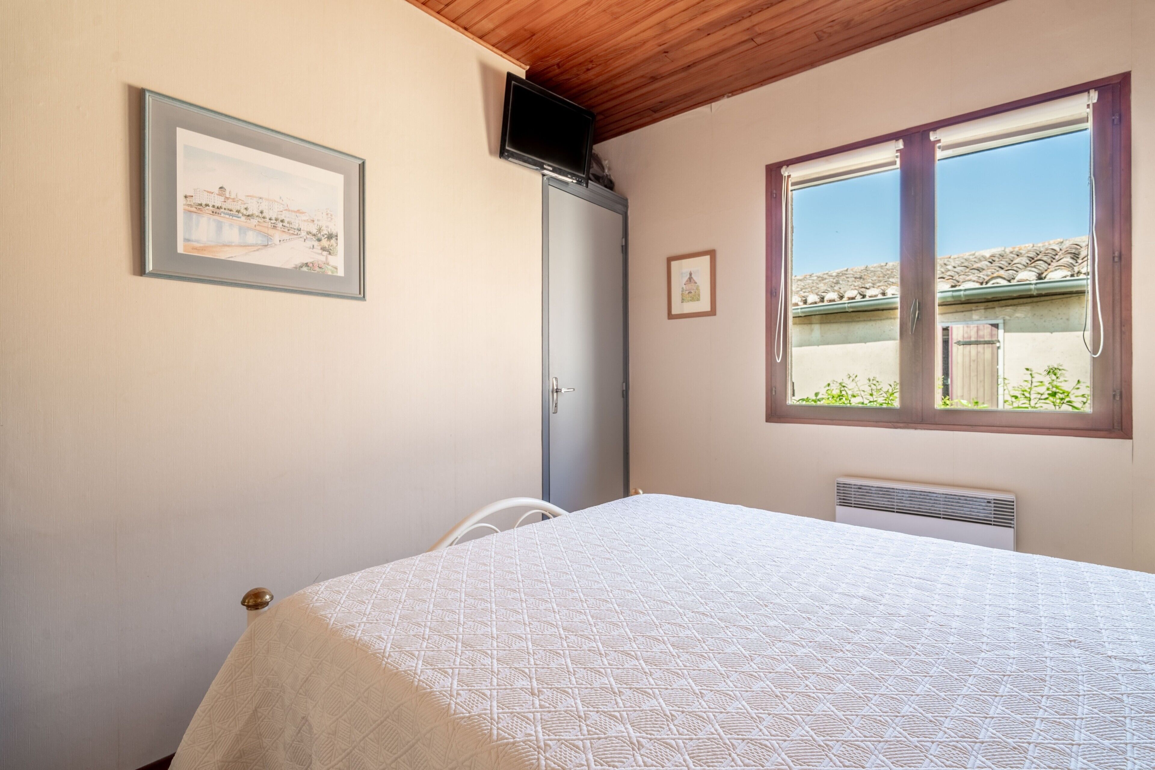 A bedroom with a bed, a window with a view, a framed picture on the wall, and a television mounted on the wall.
