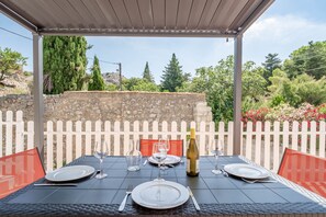 Un espace extérieur abrité, aménagé pour les repas, avec un mur en pierre, une clôture en bois peinte en blanc et de la verdure.