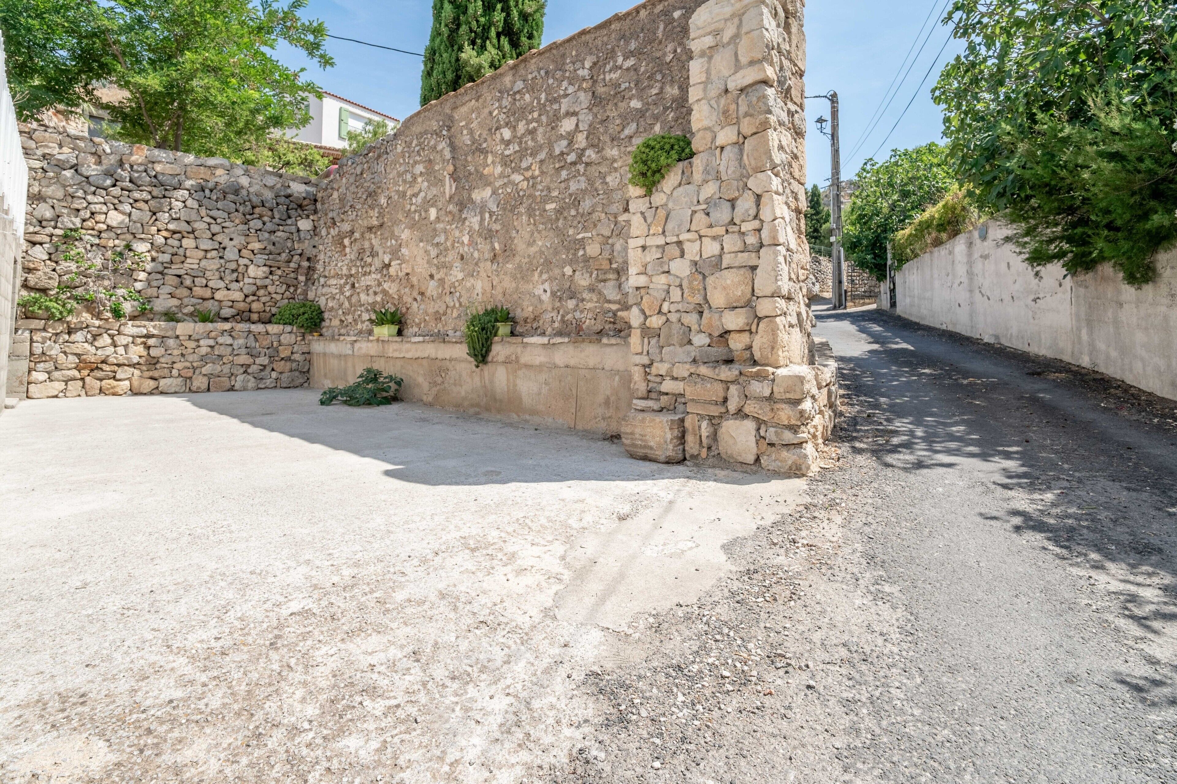 A stone wall with potted plants, a paved area, and a narrow road.