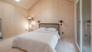 Quarto com teto inclinado, cama com roupa de cama branca, duas luminárias de cabeceira e uma janela grande.
