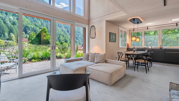 Ampla sala de estar com uma grande porta de vidro deslizante, um sofá, uma mesa de jantar e uma cozinha com vista para a floresta.