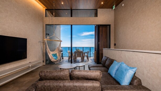 A modern living room with a large flat-screen TV, a brown leather sofa, a hanging chair, and a balcony with a view of the sea. - nagisa awajishima (Awaji)