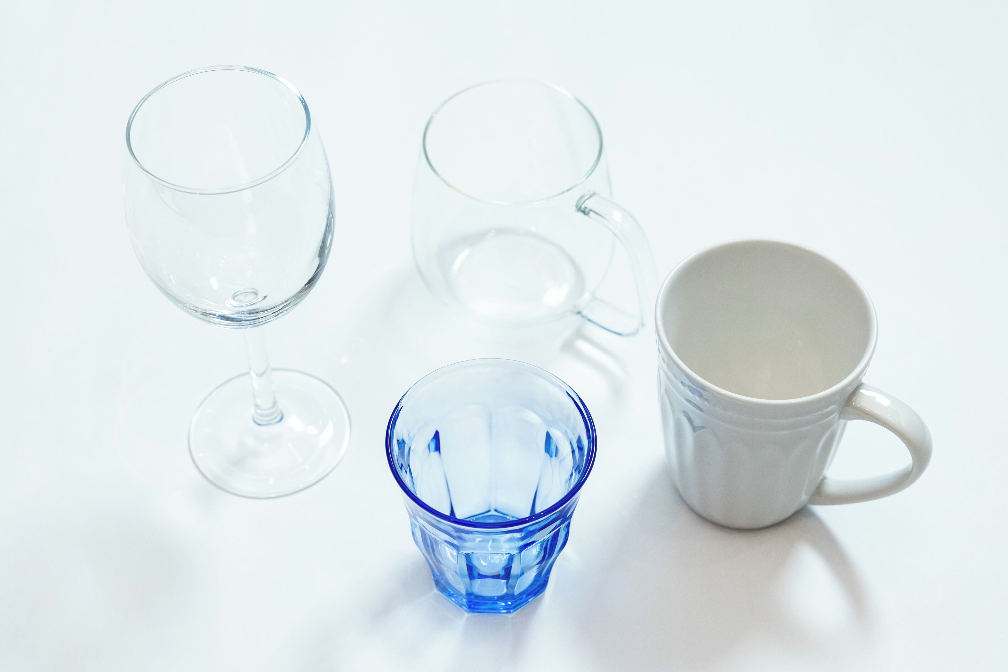 A white table with four glass items: two clear glasses, one blue glass, and one white mug.