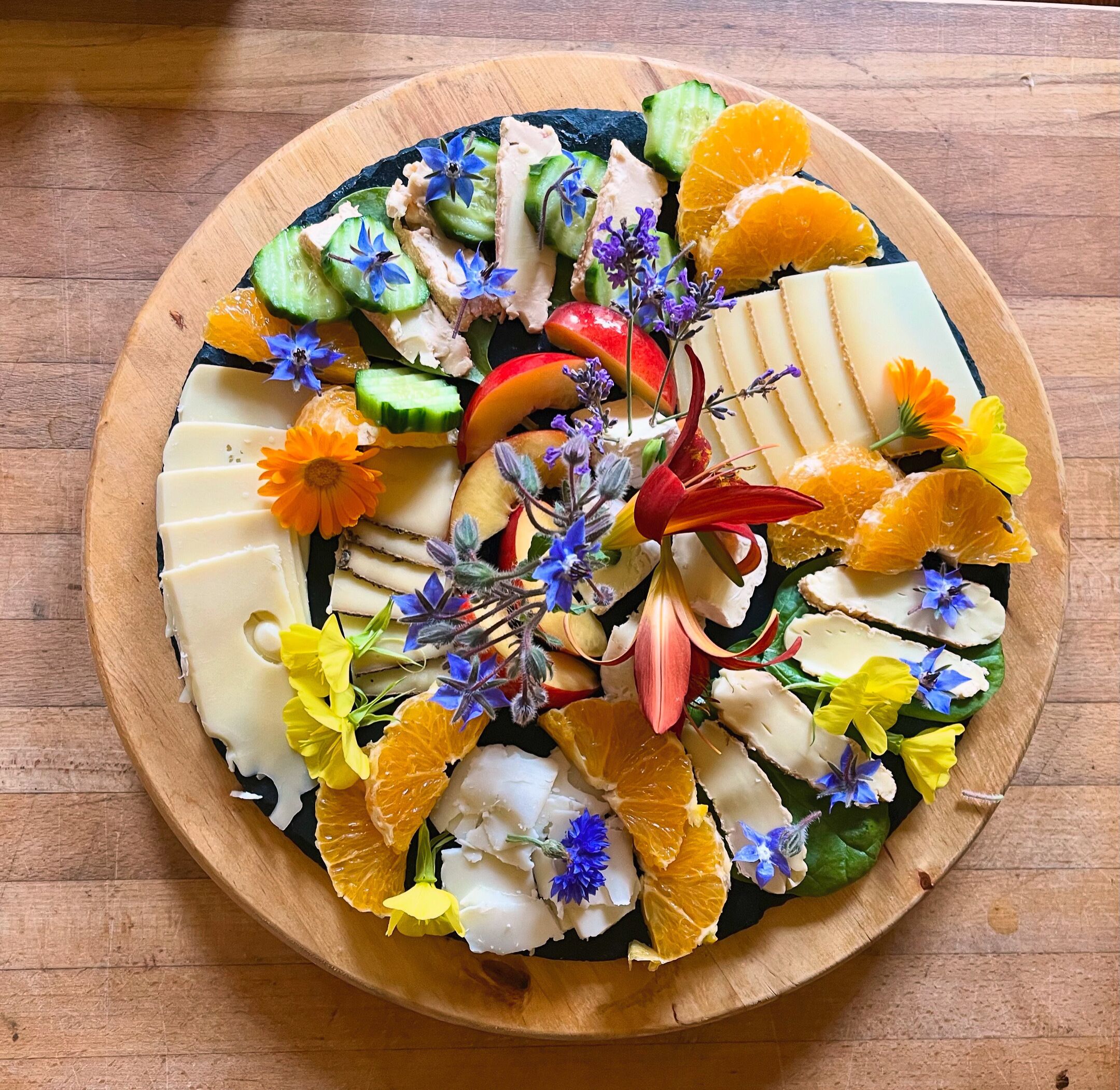 En rund træplade med et udvalg af frugt, grøntsager og blomster arrangeret på den.