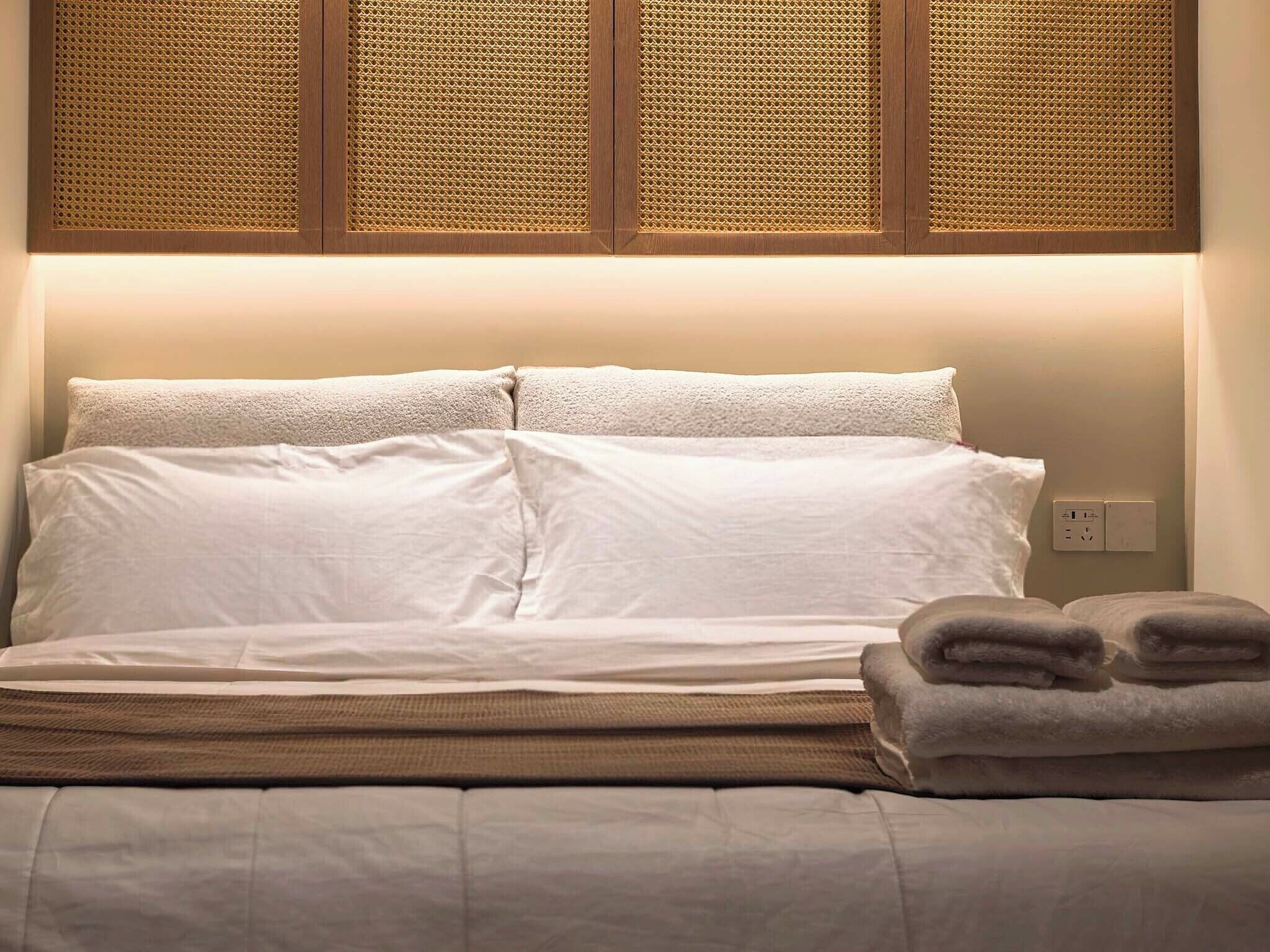 A neatly made bed with white linens, a headboard with a woven pattern, and a wall socket.