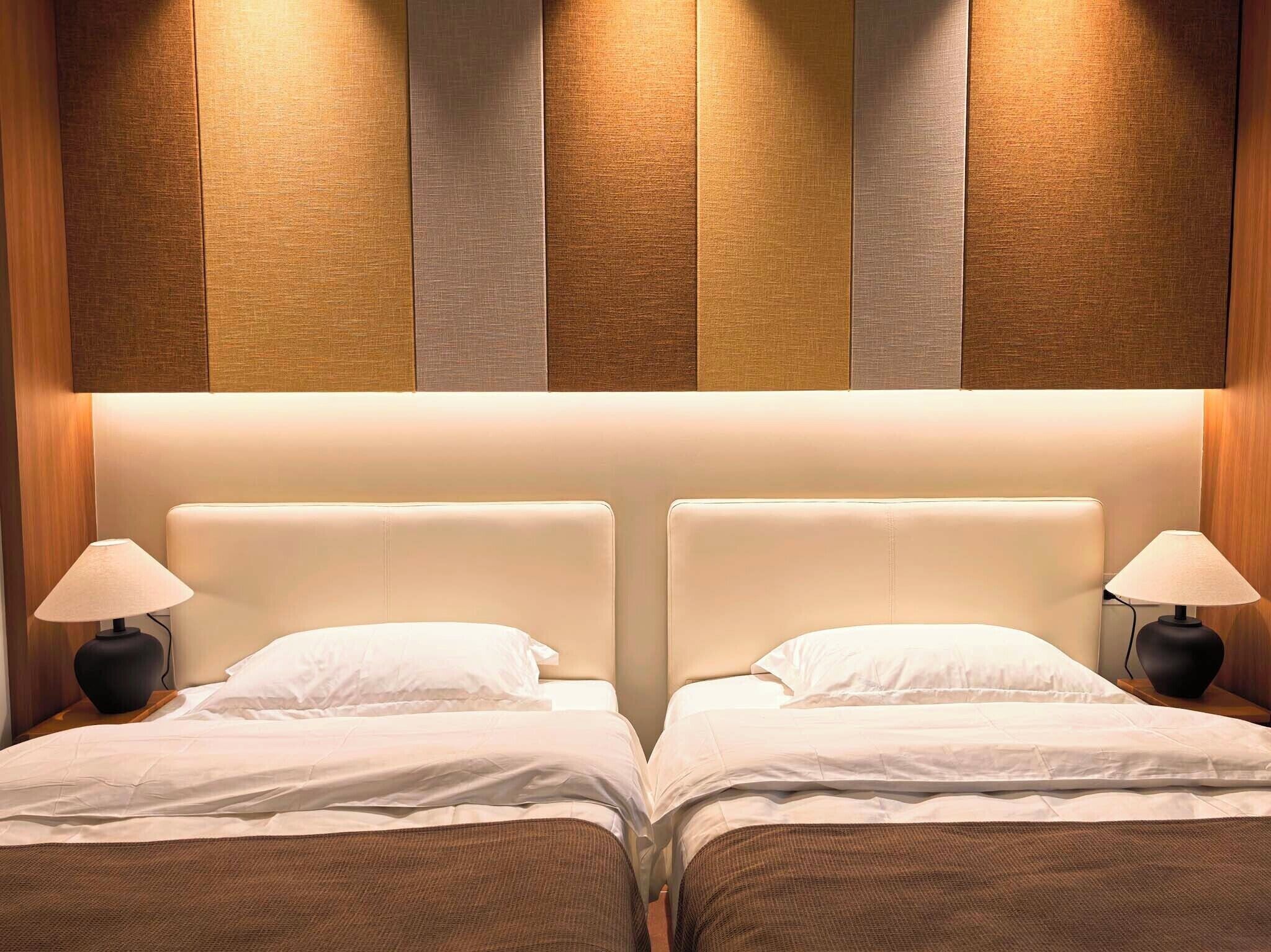 Habitación de hotel con dos camas, cada una con cabecera, lámparas de noche y una decoración en tonos neutros.