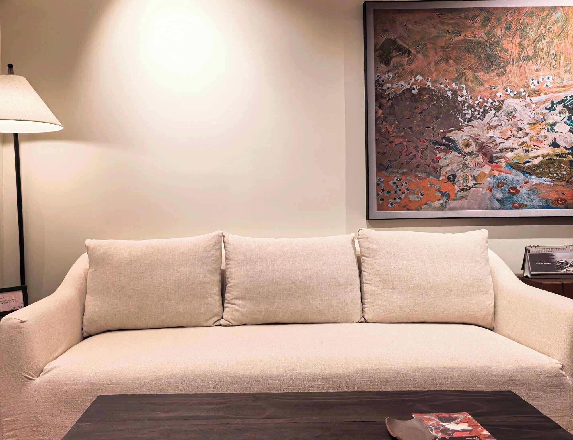 Un canapé beige avec deux coussins, une table basse en bois foncé, une lampe et un grand tableau abstrait accroché au mur.