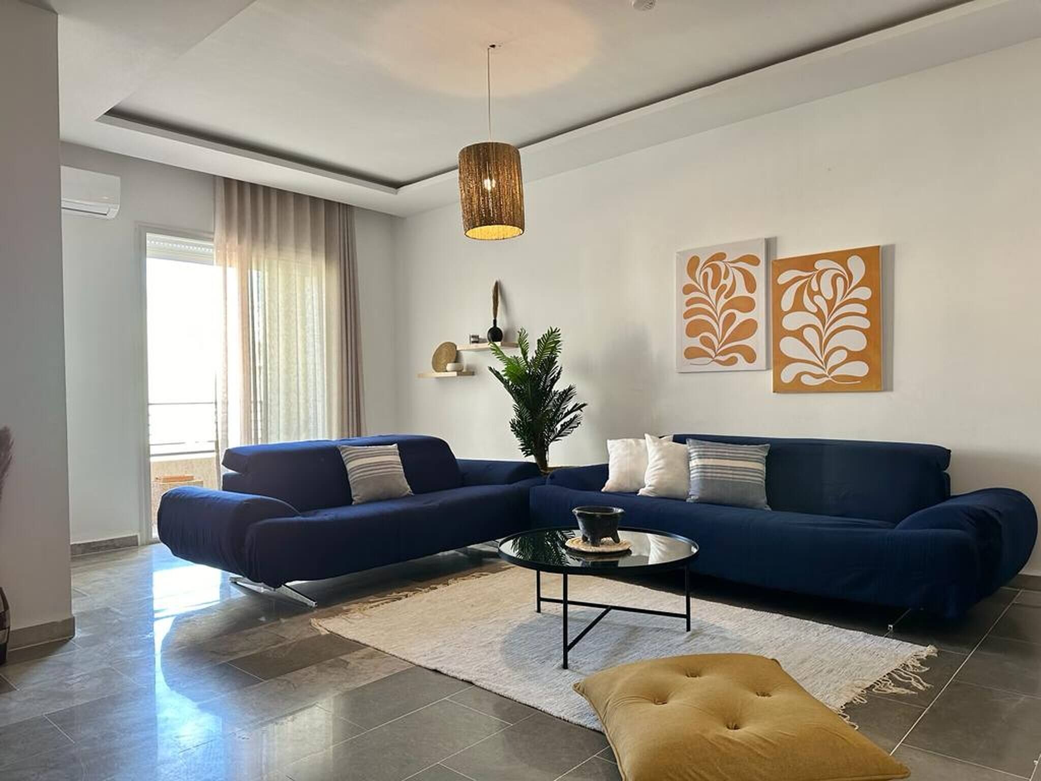 Ein modernes Wohnzimmer mit einem blauen Sofa, einem runden Couchtisch, einer hängenden Leuchte und zwei Wandbildern.